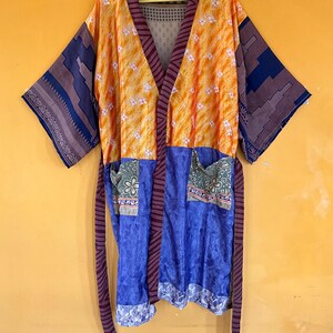 Può includere: Una colorata vestaglia kimono con un design patchwork. La vestaglia presenta pannelli arancioni, blu e viola con motivi floreali e geometrici. Ha maniche lunghe e una cintura abbinata. La vestaglia è appesa a una gruccia gialla su sfondo giallo.