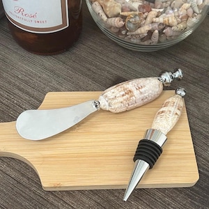 May include: A cheese knife and wine bottle stopper set. The knife and stopper handles are made from seashells. The knife has a stainless steel blade. The stopper has a black rubber seal and a silver-colored metal base.