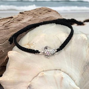 May include: Black braided bracelet with a silver turtle charm. The bracelet is displayed on a white seashell with a beach background. The turtle charm has a detailed design.