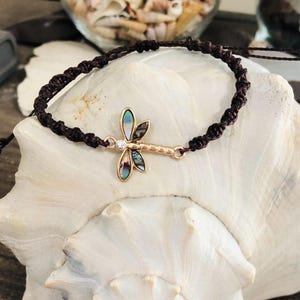 May include: A brown macrame bracelet featuring a gold-toned dragonfly charm. The dragonfly has iridescent abalone wings and a small clear crystal accent. The bracelet is displayed on a white seashell, showcasing a bohemian style.