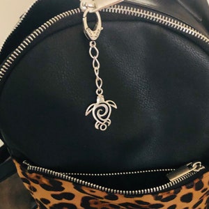 May include: A silver-colored turtle charm hanging from a black backpack. The charm is attached to a chain and a clasp. The backpack has a zipper and a leopard print pocket.