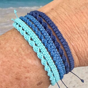 May include: A collection of six macrame bracelets in shades of blue, ranging from light turquoise to deep navy. Each bracelet is intricately knotted and features adjustable ties. The bracelets are displayed on a person's arm, with a beach scene in the background.