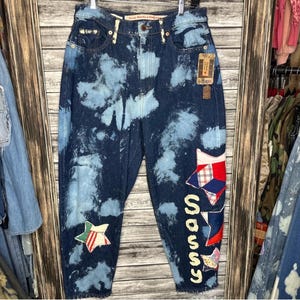 May include: Dark wash denim jeans with a light blue cloud wash effect. The jeans feature patchwork star and geometric designs, with the word "sassss" vertically down the side. A vintage style tag is attached.
