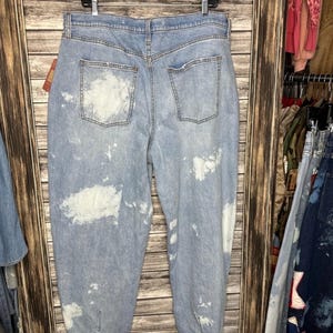 May include: Light blue denim jeans with white paint splatter details. The jeans have a high waist, two back pockets, and a relaxed fit. The jeans are hanging on a wooden background.
