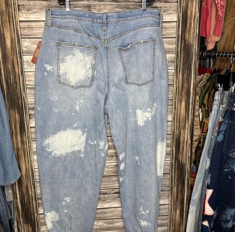 May include: Light blue denim jeans with white paint splatter details. The jeans have a high waist, two back pockets, and a relaxed fit. The jeans are hanging on a wooden background.