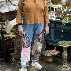 May include: A woman wearing a brown long-sleeve shirt with blue patches and pockets, paired with blue jeans featuring floral embroidery and patchwork. She is wearing white slip-on shoes. The background includes antique furniture and decor.