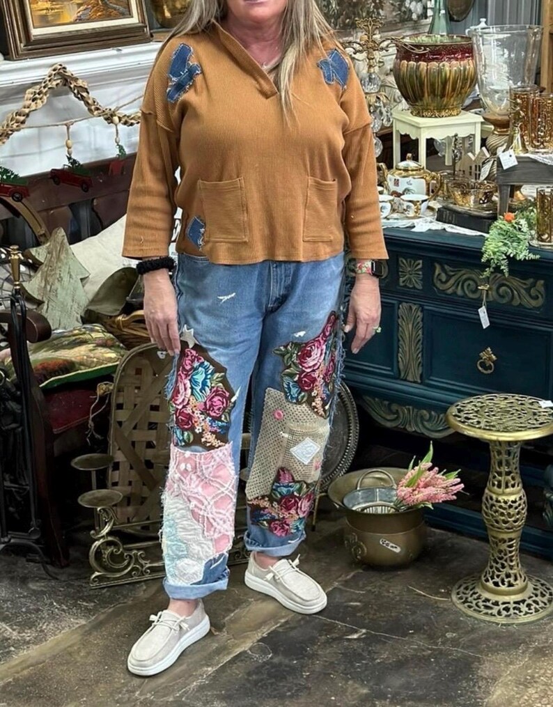 May include: A woman wearing a brown long-sleeve shirt with blue patches and pockets, paired with blue jeans featuring floral embroidery and patchwork. She is wearing white slip-on shoes. The background includes antique furniture and decor.