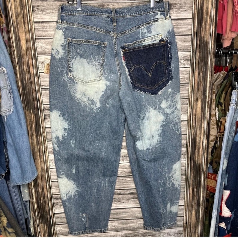 Peut inclure: Jean en denim bleu clair avec des &eacute;claboussures blanches semblables &agrave; de l'eau de Javel. Le jean pr&eacute;sente une poche plaqu&eacute;e bleu fonc&eacute; avec le logo Levi's. La vue arri&egrave;re du jean est pr&eacute;sent&eacute;e, soulignant le design unique et l'aspect vieilli.