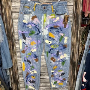 May include: Light blue denim jeans with a colorful abstract paint splatter design. The paint colors include purple, yellow, green, and brown. The jeans have a classic five-pocket design and a straight leg fit.