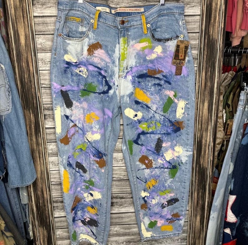 May include: Light blue denim jeans with a colorful abstract paint splatter design. The paint colors include purple, yellow, green, and brown. The jeans have a classic five-pocket design and a straight leg fit.