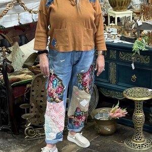 May include: A person wearing a brown long-sleeved top with blue patches and pockets, and blue jeans with floral embroidery and patchwork. They are wearing white slip-on shoes. The background features antique furniture and decor.