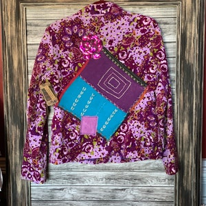 May include: A purple floral print blazer with a decorative patch. The blazer has a sequined flower detail and is hanging on a hanger. The patch is made of various colors and textures.