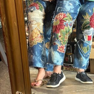 May include: Blue denim jeans with floral and lace patchwork. The jeans have a light blue tie-dye effect and are worn with sandals and trainers. The patchwork includes red, pink, and green floral designs.