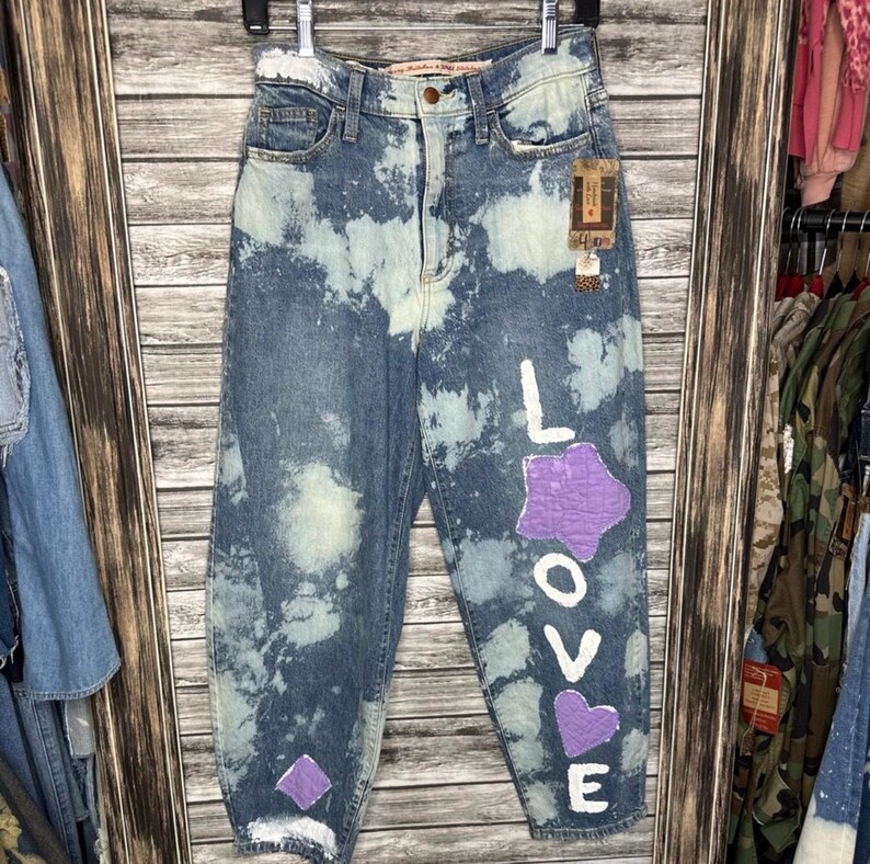 May include: Blue denim jeans with a bleached pattern and the word "LOVE" painted in white with purple accents. The jeans have a high waist and a relaxed fit, with a tag attached.