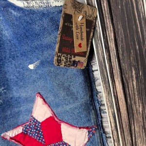 May include: Close-up of blue denim fabric with a red and blue star applique. A brown tag with the text "Handmade with Love" and "Sassy Wild Stitches" is attached to the denim.