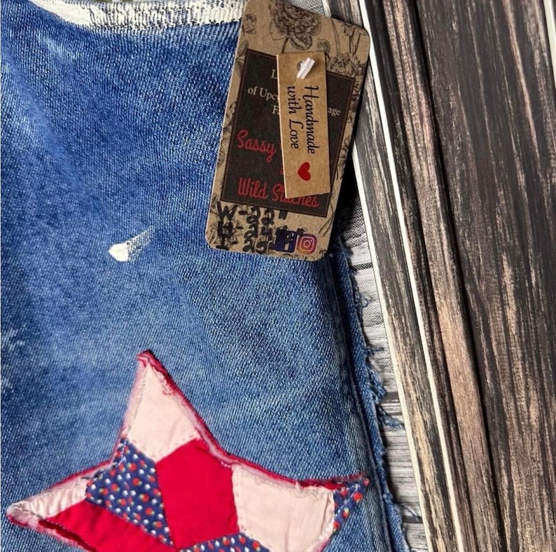 May include: Close-up of blue denim fabric with a red and blue star applique. A brown tag with the text "Handmade with Love" and "Sassy Wild Stitches" is attached to the denim.