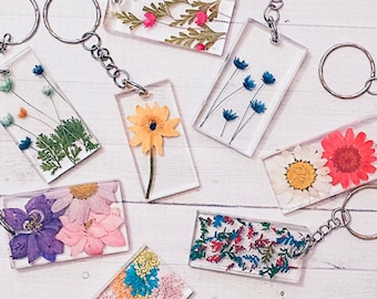 May include: A collection of rectangular resin keychains with various dried flowers embedded inside. The keychains feature a variety of colorful flowers, including yellow, pink, blue, and white, and are attached to silver key rings. The background is a light wood surface.
