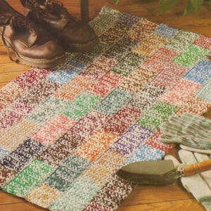 May include: A multi-colored crocheted rug with a patchwork design. The rug is made with different shades of brown, green, pink, and blue yarn.