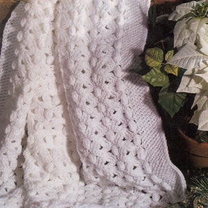 May include: A white crocheted blanket with a textured, raised pattern. The blanket is folded and draped over a decorative box with a gold ribbon. The blanket is surrounded by greenery and white poinsettia flowers.