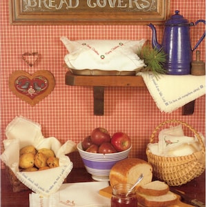 May include: A rustic kitchen scene with a wooden sign reading "BREAD COVERS." Features include a blue enamel teapot, bread, apples, and baskets. Decorative linens with embroidered text and a heart-shaped wall hanging add charm.