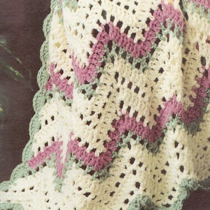 May include: A crocheted blanket featuring a chevron pattern in cream, dusty rose, and sage green. The textile has an openwork stitch design and a scalloped edge border in sage green, showcasing a soft, textured yarn construction throughout.