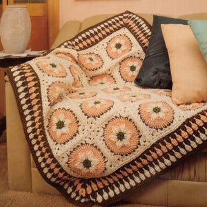 May include: A handmade crochet blanket with a floral pattern in cream, peach, brown, and green. The blanket is draped over a sofa, showcasing its intricate design and textured stitches. The border features a scalloped edge.