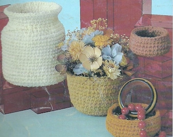 May include: A set of three crocheted baskets in beige and brown. The largest basket is filled with artificial flowers and is sitting on a brown surface. The smallest basket is sitting on a blue surface and contains a bracelet.