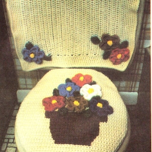 Op de afbeelding: Een gehaakte toiletbrilhoes en tankhoes met een bloemenmotief. De brilhoes heeft een boeket gehaakte bloemen in een bruine pot. De tankhoes heeft een enkele gehaakte bloem met een bruin hart en rode en witte bloemblaadjes.