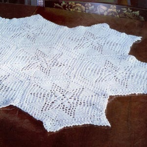 Może przedstawiać: Biały, ręcznie robiony koc z szydełka o szczegółowym ażurowym wzorze. Koc ma falisty brzeg i teksturowaną powierzchnię. Złożony wzór zawiera powtarzające się kształty geometryczne, tworząc delikatny i przewiewny wygląd. Ten koc jest idealny, aby dodać odrobinę uroku ręcznie robionego do każdego pomieszczenia.