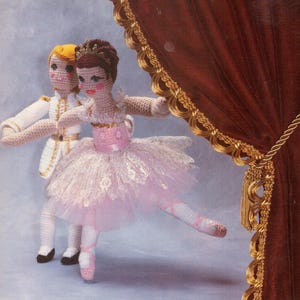 May include: Crocheted ballerina and prince dolls in a performance pose, with a red curtain in the background. The ballerina wears a pink tutu and a crown. The prince is dressed in a white suit with gold trim.