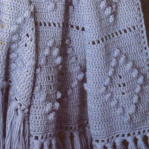 May include: A light blue crochet blanket featuring a repeating diamond pattern created with raised bobble stitches. The fabric includes sections of openwork mesh and is finished with long, thick fringe tassels along the bottom edge.