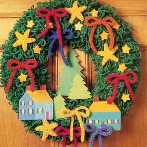 May include: A green yarn wreath decorated with yellow fabric stars, red, orange, blue, and yellow yarn bows, and two flat wooden houses in blue and yellow. The center features two green paper pine trees, creating a festive, layered wall hanging.