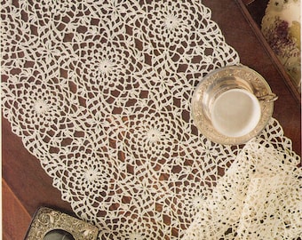 May include: A white crocheted table runner with a delicate floral pattern. The runner is laid out on a wooden table with a silver teacup and saucer, a small decorative box, and a bouquet of pink and white roses.