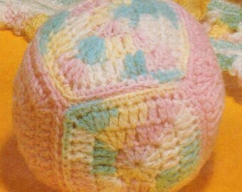 Puede incluir: Un juguete de pelota de crochet en rosa, amarillo, azul y blanco. La pelota está hecha de varios cuadrados de diferentes colores.