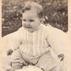 Op de afbeelding: Een baby draagt een wit gebreid vest met knopen. De baby zit op een wit deken en heeft een gelukkige uitdrukking op zijn gezicht.