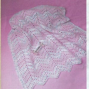 May include: A pastel pink and blue crocheted baby blanket with a scalloped edge. A silver rattle rests on the blanket. The blanket has a delicate, open weave pattern.