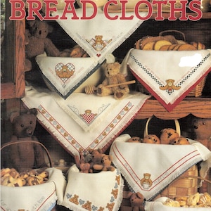 May include: A collection of embroidered bread cloths featuring teddy bears and heartwarming sayings. The cloths are displayed on a wooden shelf with baskets of bread and muffins.
