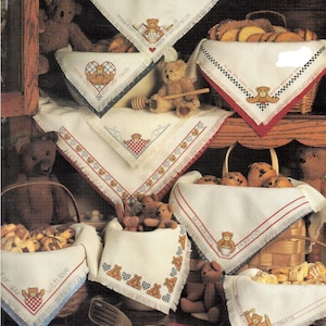May include: A collection of off-white fabric basket liners featuring cross-stitched teddy bear motifs, red and blue checkered patterns, and embroidered text including "made from scratch," "with love," and "blueberry muffins" with fringed edges.