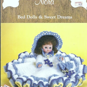 May include: A crochet doll with a white dress, blue hat, and a matching blue crochet purse. The doll is sitting on a white and blue crocheted bedspread. The pattern is called "Nora Bed Dolls & Sweet Dreams".
