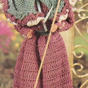 Puede incluir: Muñeca vestida con un atuendo de ganchillo hecho a mano. El atuendo incluye un vestido largo malva, una capa verde con ribete blanco y un sombrero a juego con plumas y una rosa. La muñeca sostiene un palo de madera y una pequeña bolsa.
