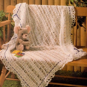 May include: A white, open-weave crochet baby blanket with a scalloped edge and purple ribbon accents. A plush gray teddy bear and colorful toy are on the blanket, which is draped over a wooden bench. The blanket is approximately 100 x 100 cm.