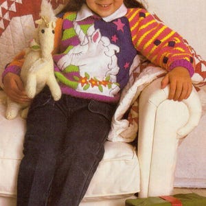 Può includere: Una bambina con un maglione di lana colorato con un disegno di unicorno è seduta su una poltrona bianca tenendo un unicorno di peluche bianco. Indossa jeans blu e calzini bianchi. Una scatola regalo verde avvolta con un nastro rosso e un disegno di fiocco di neve è sul pavimento davanti alla poltrona.