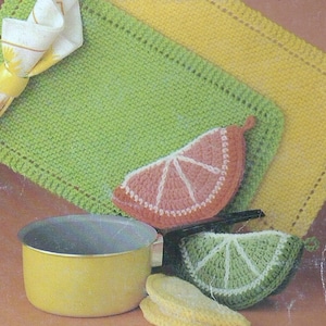 Op de afbeelding: Drie gebreide pottenlapjes in de vorm van citrusvruchtenplakjes. De pottenlapjes zijn groen, geel en roze. De pottenlapjes liggen op een bruin oppervlak met een gele pot.