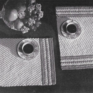 May include: Two crocheted placemats with a textured design. The placemats are white with a light gray pattern and have a dark gray border. A white saucer with a cup and a dark liquid is on top of one of the placemats.