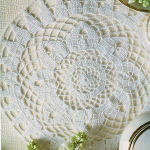 May include: A close-up of a white, crocheted doily with a floral design. The doily is circular and has a delicate, openwork pattern. The background is a light beige color, and there are sprigs of white flowers and a decorative plate.