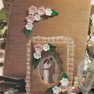 May include: A brown velvet photo album with a crocheted flower design in pink and white. The album is decorated with green leaves. The album is closed and sitting on a table with a lace tablecloth. There is a smaller photo frame with a picture of a couple in it. The photo frame is also decorated with crocheted flowers and leaves.