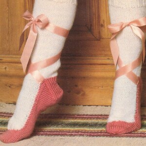 May include: White knitted socks with pink ballet slipper toes and pink satin ribbon bows. The socks are worn on a pair of legs.