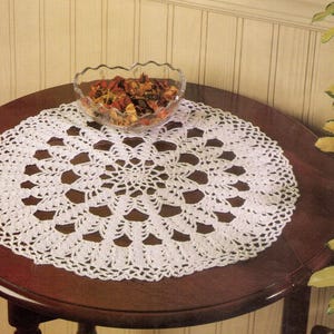 May include: A white, crocheted doily with a detailed openwork pattern sits on a dark brown, round table. A clear glass bowl filled with dried potpourri rests on the doily. The table is in front of a light-colored wall.