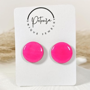 May include: A pair of bright pink, round stud earrings. The earrings are displayed on a white card with the brand name "Petruza Unique Jewelry" printed on it. The earrings are on a white, decorative plate.