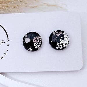 Puede incluir: Un par de pendientes de botón redondos con una superficie negra brillante y copos de lámina metálica plateada incrustados. Los pendientes están sujetos a una tarjeta de presentación blanca con el texto Petruža UNIQUE JEWELRY.